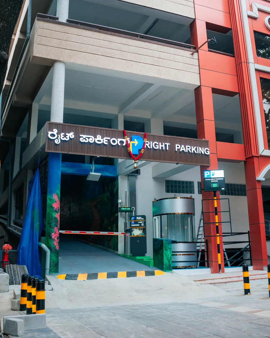 Right Parking at Gandhi Bazaar — multi-level street-front facility with illuminated bilingual signage, boom barrier lane and a live available-spaces display
